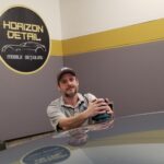 The owner posing over a hood he just sanded and refinished. Smiling holding a sander with his logo behind him and reflecting in the hood